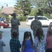 Two U.S. Army soldiers in camouflage uniforms are speaking to a group of young children outdoors on a paved surface. The setting appears to be a community or school event where military personnel are engaging with children, possibly for an educational or outreach visit. The scene takes place in a small town or suburban area with buildings, trees, and parked vehicles visible in the background.