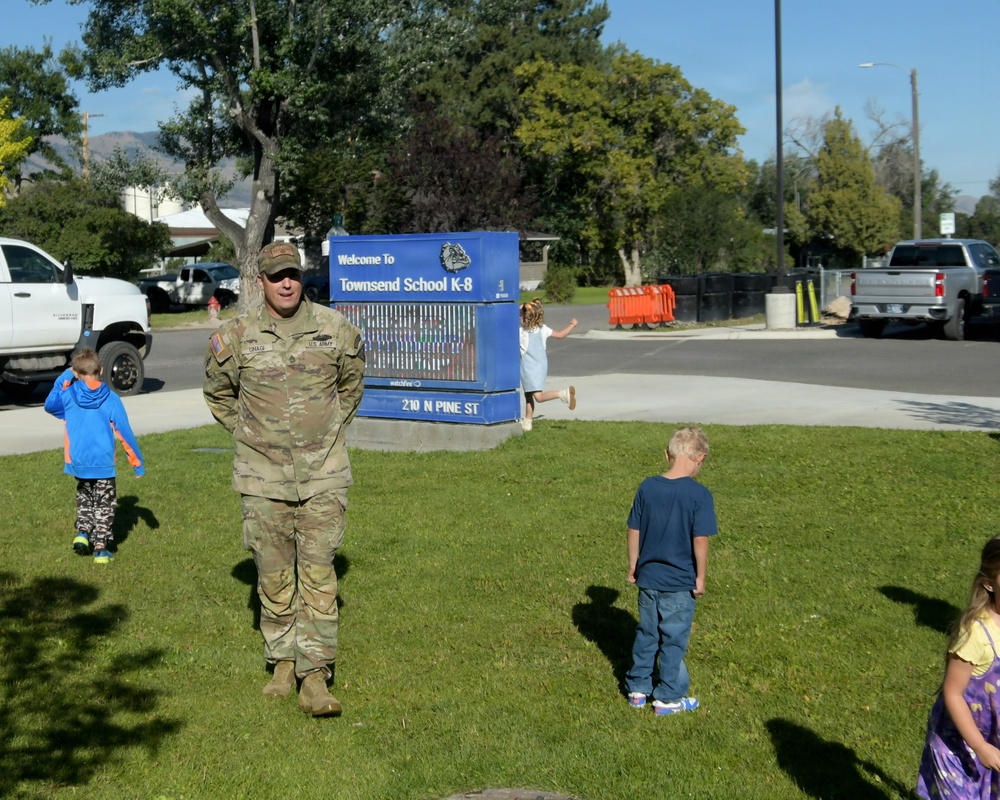 A U.S. Army soldier in OCP (Operational Camouflage Pattern) uniform stands on a grass lawn in front of Townsend School K-8, located at 210 N Pine St. Several children are visible around him on the school grounds, suggesting a school visit or community engagement event. The setting is a sunny day with trees, parked vehicles, and a blue school marquee sign visible in the background.