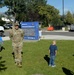 A U.S. Army soldier in OCP (Operational Camouflage Pattern) uniform stands on a grass lawn in front of Townsend School K-8, located at 210 N Pine St. Several children are visible around him on the school grounds, suggesting a school visit or community engagement event. The setting is a sunny day with trees, parked vehicles, and a blue school marquee sign visible in the background.