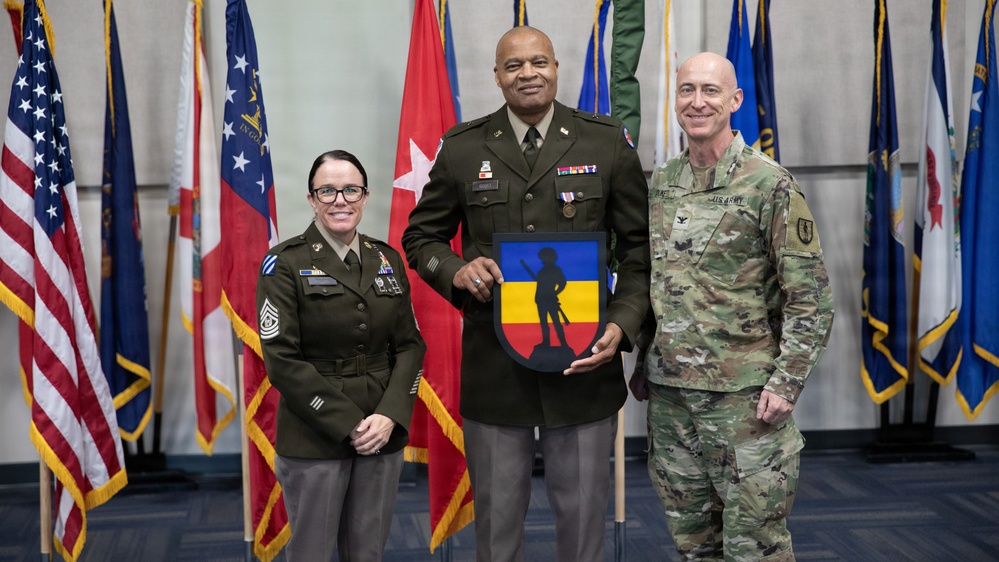 Three U.S. Army personnel pose for a formal photo in front of a display of military and state flags. The central figure, wearing Army Service Uniform (green), holds a decorative shield-shaped plaque featuring a soldier silhouette on a blue and yellow background. The occasion appears to be a military ceremony or award presentation.