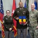 Three U.S. Army personnel pose for a formal photo in front of a display of military and state flags. The central figure, wearing Army Service Uniform (green), holds a decorative shield-shaped plaque featuring a soldier silhouette on a blue and yellow background. The occasion appears to be a military ceremony or award presentation.