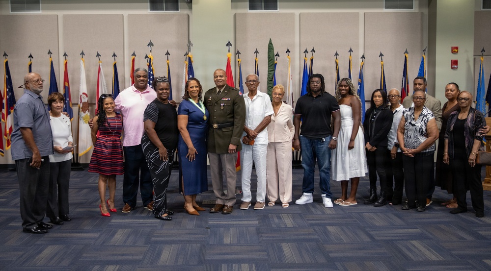A group of approximately 18-20 individuals pose for a formal group photograph in what appears to be a military ceremony hall. A central figure in U.S. Army dress uniform with medals is flanked by family members and guests, suggesting a military retirement, promotion, or award ceremony. The setting features numerous military unit and state flags displayed in the background.