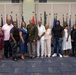 A group of approximately 18-20 individuals pose for a formal group photograph in what appears to be a military ceremony hall. A central figure in U.S. Army dress uniform with medals is flanked by family members and guests, suggesting a military retirement, promotion, or award ceremony. The setting features numerous military unit and state flags displayed in the background.
