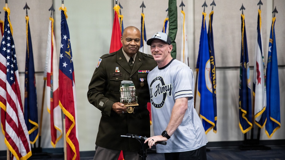 Two individuals pose together in what appears to be a formal military ceremony or award presentation. A uniformed U.S. Army officer wearing the name tag 'SCOTT' holds a trophy or commemorative item, standing alongside a civilian or off-duty military member wearing a '2021 All Army Basketball' shirt. Multiple U.S. state and military flags are displayed in the background.