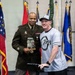 Two individuals pose together in what appears to be a formal military ceremony or award presentation. A uniformed U.S. Army officer wearing the name tag 'SCOTT' holds a trophy or commemorative item, standing alongside a civilian or off-duty military member wearing a '2021 All Army Basketball' shirt. Multiple U.S. state and military flags are displayed in the background.