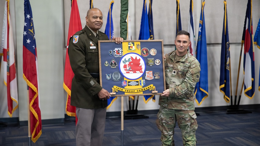 Two U.S. Army personnel are posing together while holding a shadow box display board labeled '78th Troop Command,' featuring a red boar insignia and multiple unit coins and patches. The individual on the left is wearing the Army Green Service Uniform, while the individual on the right is in Army Combat Uniform (ACU/OCP). The ceremony appears to be a formal military presentation or award event, set against a backdrop of multiple state and military flags.