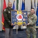 Two U.S. Army personnel are posing together while holding a shadow box display board labeled '78th Troop Command,' featuring a red boar insignia and multiple unit coins and patches. The individual on the left is wearing the Army Green Service Uniform, while the individual on the right is in Army Combat Uniform (ACU/OCP). The ceremony appears to be a formal military presentation or award event, set against a backdrop of multiple state and military flags.