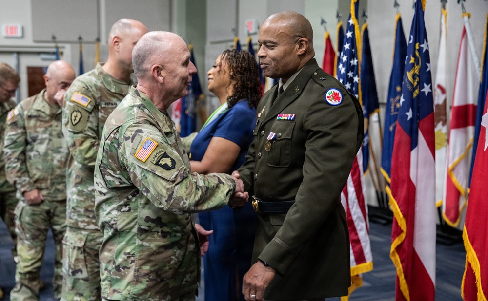 A formal U.S. Army military ceremony is taking place in an indoor venue with multiple flags displayed in the background. Two soldiers are shaking hands at the center of the image — one in Army Combat Uniform (ACU/OCP camouflage) and one in the Army Green Service Uniform. Additional military personnel and a civilian woman are visible in the background.
