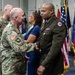 A formal U.S. Army military ceremony is taking place in an indoor venue with multiple flags displayed in the background. Two soldiers are shaking hands at the center of the image — one in Army Combat Uniform (ACU/OCP camouflage) and one in the Army Green Service Uniform. Additional military personnel and a civilian woman are visible in the background.