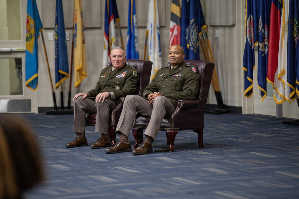 Two military officers in olive green dress uniforms are seated in leather wingback chairs during what appears to be a formal military ceremony or event. They are positioned in front of a display of numerous military and state flags, suggesting a significant official occasion. Both individuals wear rank insignia and service ribbons on their uniforms.