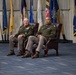 Two military officers in olive green dress uniforms are seated in leather wingback chairs during what appears to be a formal military ceremony or event. They are positioned in front of a display of numerous military and state flags, suggesting a significant official occasion. Both individuals wear rank insignia and service ribbons on their uniforms.