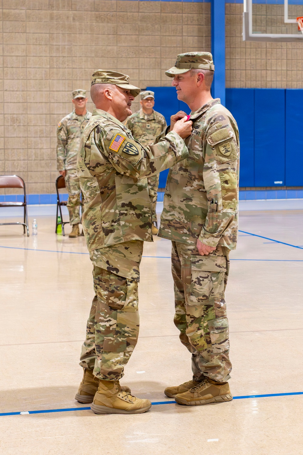 A U.S. Army medal pinning ceremony taking place inside a gymnasium or indoor sports facility. One soldier is pinning a medal or decoration onto the chest of another soldier, both wearing Operational Camouflage Pattern (OCP) uniforms. Additional soldiers stand at attention in the background as witnesses to the ceremony.