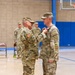 A U.S. Army medal pinning ceremony taking place inside a gymnasium or indoor sports facility. One soldier is pinning a medal or decoration onto the chest of another soldier, both wearing Operational Camouflage Pattern (OCP) uniforms. Additional soldiers stand at attention in the background as witnesses to the ceremony.