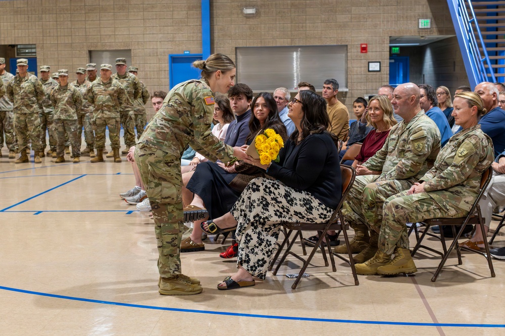 A military ceremony taking place in an indoor gymnasium or armory, where a female soldier in Army Combat Uniform is presenting a bouquet of yellow roses to a seated civilian woman. Rows of uniformed soldiers stand at attention in the background, while civilians and additional military personnel are seated as an audience. The event appears to be a formal recognition or farewell ceremony.