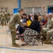 A military ceremony taking place in an indoor gymnasium or armory, where a female soldier in Army Combat Uniform is presenting a bouquet of yellow roses to a seated civilian woman. Rows of uniformed soldiers stand at attention in the background, while civilians and additional military personnel are seated as an audience. The event appears to be a formal recognition or farewell ceremony.