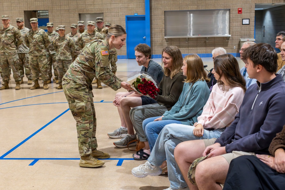 A female U.S. Army soldier in OCP (Operational Camouflage Pattern) uniform is presenting a bouquet of red roses to a seated civilian woman during what appears to be a military ceremony in a gymnasium. A formation of soldiers stands at attention in the background. The setting and gesture suggest a farewell, deployment, or recognition ceremony.