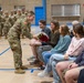 A female U.S. Army soldier in OCP (Operational Camouflage Pattern) uniform is presenting a bouquet of red roses to a seated civilian woman during what appears to be a military ceremony in a gymnasium. A formation of soldiers stands at attention in the background. The setting and gesture suggest a farewell, deployment, or recognition ceremony.