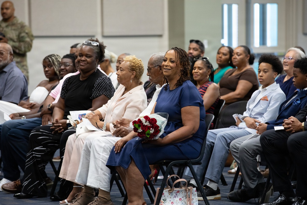 A group of people are seated in rows of chairs at what appears to be a formal ceremony or event, likely a military promotion, graduation, or retirement ceremony. A woman in a blue dress prominently holds a bouquet of red roses, suggesting she may be the honoree or a close family member of the honoree. A person in military camouflage uniform is partially visible in the background.