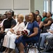 A group of people are seated in rows of chairs at what appears to be a formal ceremony or event, likely a military promotion, graduation, or retirement ceremony. A woman in a blue dress prominently holds a bouquet of red roses, suggesting she may be the honoree or a close family member of the honoree. A person in military camouflage uniform is partially visible in the background.