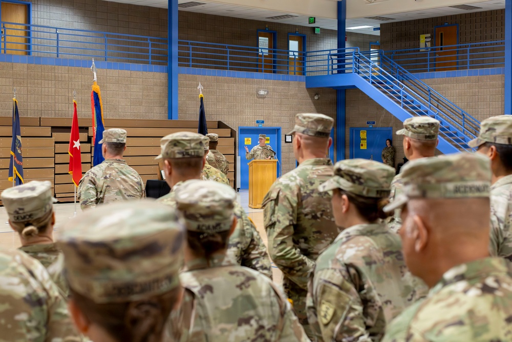 A military ceremony or briefing is taking place inside a gymnasium or multipurpose facility, with a uniformed soldier speaking at a wooden podium. A formation of soldiers in Army Combat Uniforms (ACU/OCP pattern) stands at attention facing the speaker. Several military flags, including what appears to be a general officer's flag (red with star), are displayed at the front of the room.