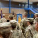 A military ceremony or briefing is taking place inside a gymnasium or multipurpose facility, with a uniformed soldier speaking at a wooden podium. A formation of soldiers in Army Combat Uniforms (ACU/OCP pattern) stands at attention facing the speaker. Several military flags, including what appears to be a general officer's flag (red with star), are displayed at the front of the room.