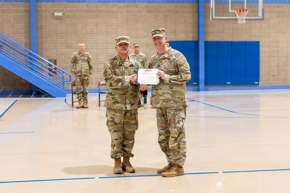 Two U.S. Army soldiers stand in the center of an indoor gymnasium posing for a photo while jointly holding a certificate or award document. The soldier on the right is wearing a medal pinned to his chest, indicating this is a military awards ceremony. Additional soldiers are visible in the background observing the presentation.