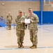 Two U.S. Army soldiers stand in the center of an indoor gymnasium posing for a photo while jointly holding a certificate or award document. The soldier on the right is wearing a medal pinned to his chest, indicating this is a military awards ceremony. Additional soldiers are visible in the background observing the presentation.