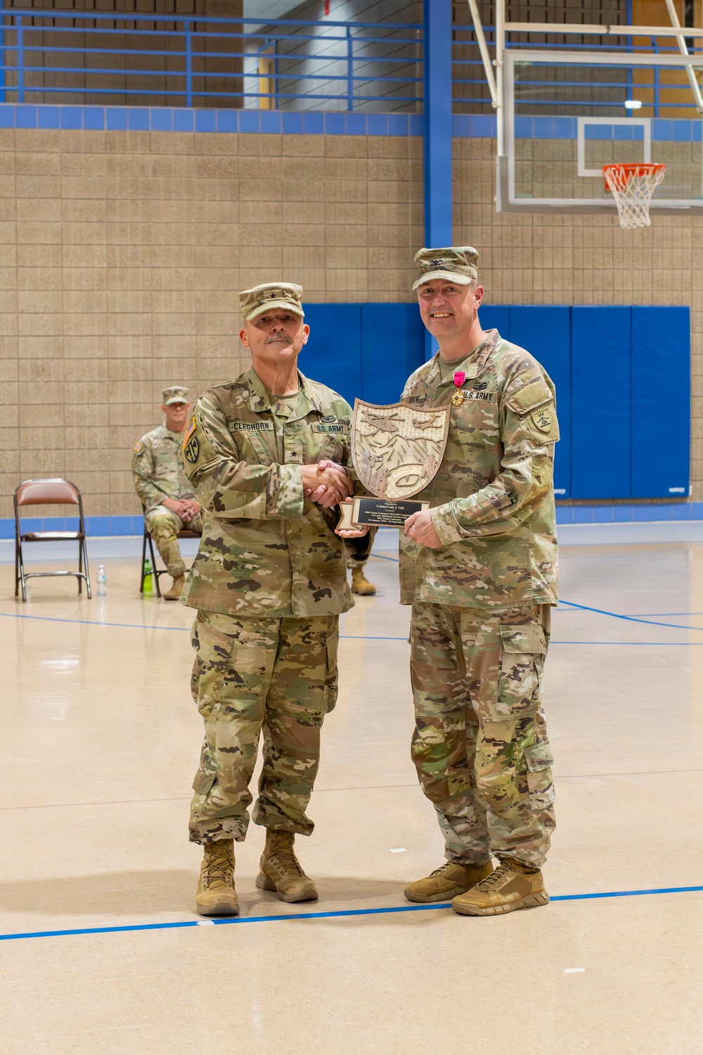 Two U.S. Army soldiers in OCP (Operational Camouflage Pattern) uniforms are posing together in a gymnasium while jointly holding a shield-shaped plaque or award. The soldier on the left, identified by his name tape as 'CLEGHORN,' appears to hold a general officer rank insignia on his cap. The soldier on the right is wearing a medal on his chest, suggesting this is an awards or recognition ceremony.