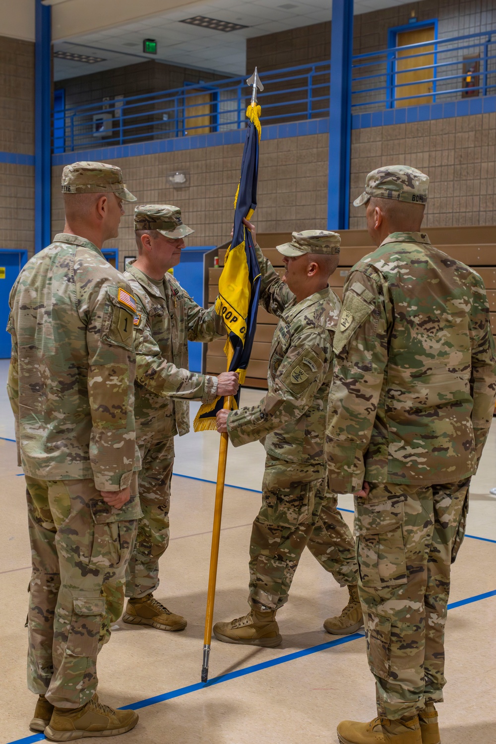 A U.S. Army change of command ceremony is taking place inside a gymnasium or indoor facility. Four soldiers in OCP (Operational Camouflage Pattern) uniforms are participating in the formal passing of a unit guidon, which is the central act of a change of command ceremony. The guidon appears to be a blue and gold flag with text reading 'TROOP C' or similar unit designation.
