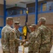 A U.S. Army change of command ceremony is taking place inside a gymnasium or indoor facility. Four soldiers in OCP (Operational Camouflage Pattern) uniforms are participating in the formal passing of a unit guidon, which is the central act of a change of command ceremony. The guidon appears to be a blue and gold flag with text reading 'TROOP C' or similar unit designation.