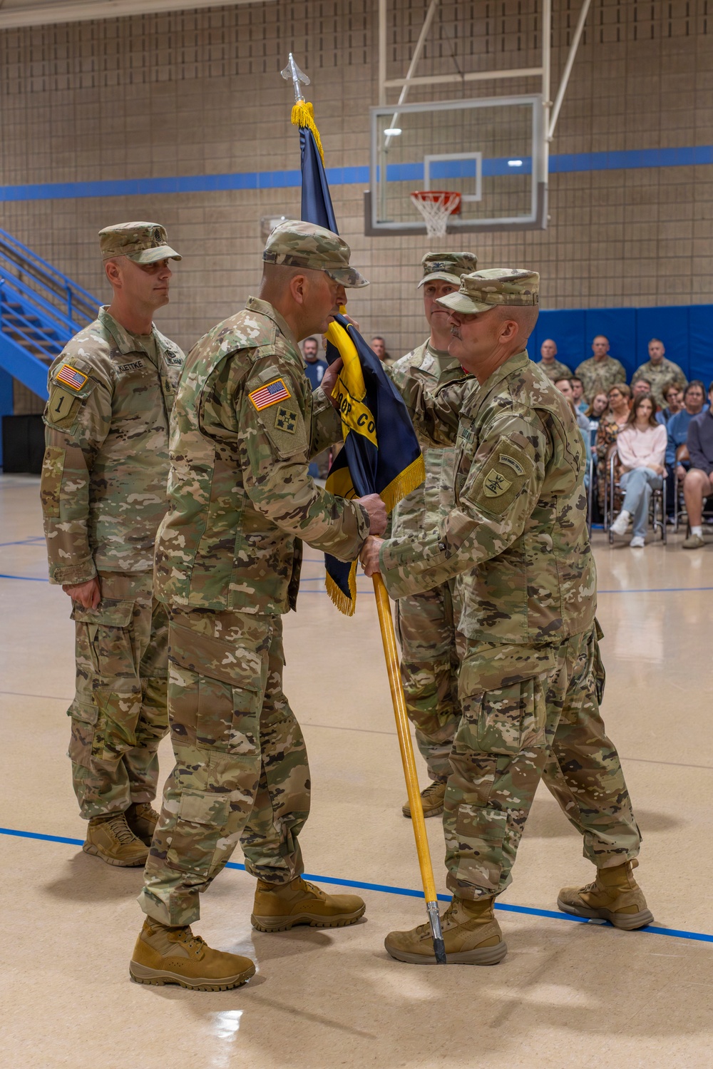 A U.S. Army change of command ceremony is taking place inside a gymnasium or armory. Two soldiers in the foreground are exchanging a unit guidon (flag), which is the central symbolic act of a change of command. Seated spectators and additional uniformed personnel observe the ceremony in the background.