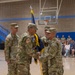 A U.S. Army change of command ceremony is taking place inside a gymnasium or armory. Two soldiers in the foreground are exchanging a unit guidon (flag), which is the central symbolic act of a change of command. Seated spectators and additional uniformed personnel observe the ceremony in the background.