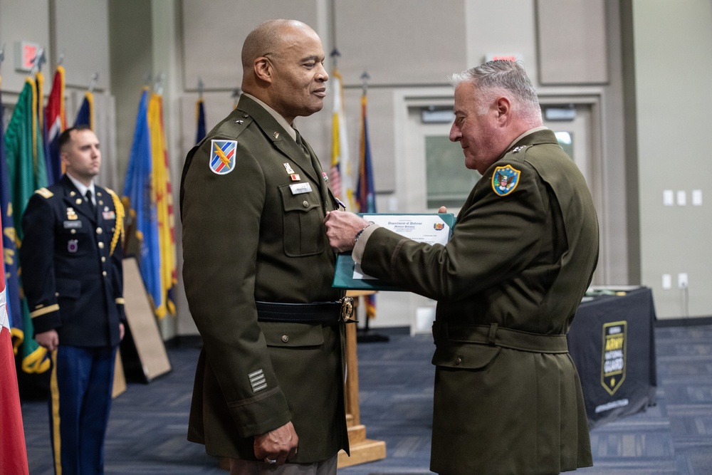 A military ceremony is taking place in which one officer is pinning a medal or award onto another officer's uniform while presenting a Department of Defense certificate. Both individuals are wearing U.S. Army green service uniforms with various insignia and patches. The event appears to be a formal awards or promotion ceremony, with color guard flags and an Iowa Army National Guard banner visible in the background.