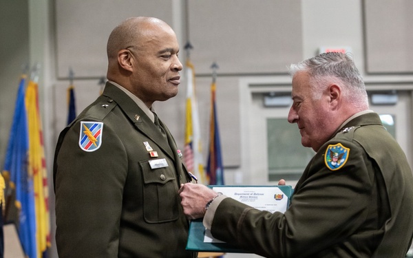 A military ceremony is taking place in which one officer is pinning a medal or award onto another officer's uniform while presenting a Department of Defense certificate. Both individuals are wearing U.S. Army green service uniforms with various insignia and patches. The event appears to be a formal awards or promotion ceremony, with color guard flags and an Iowa Army National Guard banner visible in the background.