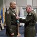 A military ceremony is taking place in which one officer is pinning a medal or award onto another officer's uniform while presenting a Department of Defense certificate. Both individuals are wearing U.S. Army green service uniforms with various insignia and patches. The event appears to be a formal awards or promotion ceremony, with color guard flags and an Iowa Army National Guard banner visible in the background.
