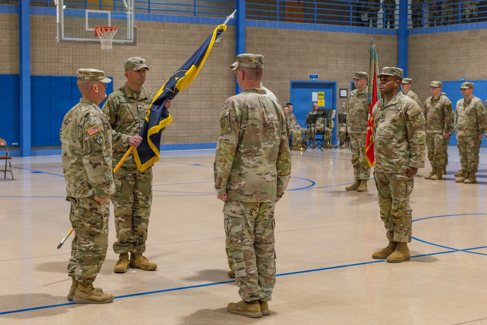 A U.S. Army change of command or activation ceremony is taking place inside a gymnasium. A central figure is holding a unit guidon/flag with blue and gold colors, which is being passed or presented during the formal military ceremony. Soldiers in Army Combat Uniforms (ACU/OCP pattern) are arranged in formation on both sides.