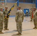 A U.S. Army change of command or activation ceremony is taking place inside a gymnasium. A central figure is holding a unit guidon/flag with blue and gold colors, which is being passed or presented during the formal military ceremony. Soldiers in Army Combat Uniforms (ACU/OCP pattern) are arranged in formation on both sides.