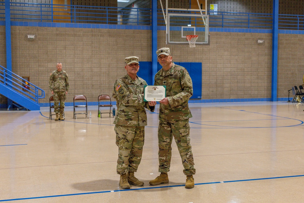 Two U.S. Army soldiers in MultiCam (OCP) camouflage uniforms are posing together while jointly holding a certificate or award document in what appears to be a military gymnasium. A third soldier stands in the background near a staircase. This appears to be a formal military awards or recognition ceremony.