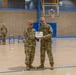 Two U.S. Army soldiers in MultiCam (OCP) camouflage uniforms are posing together while jointly holding a certificate or award document in what appears to be a military gymnasium. A third soldier stands in the background near a staircase. This appears to be a formal military awards or recognition ceremony.