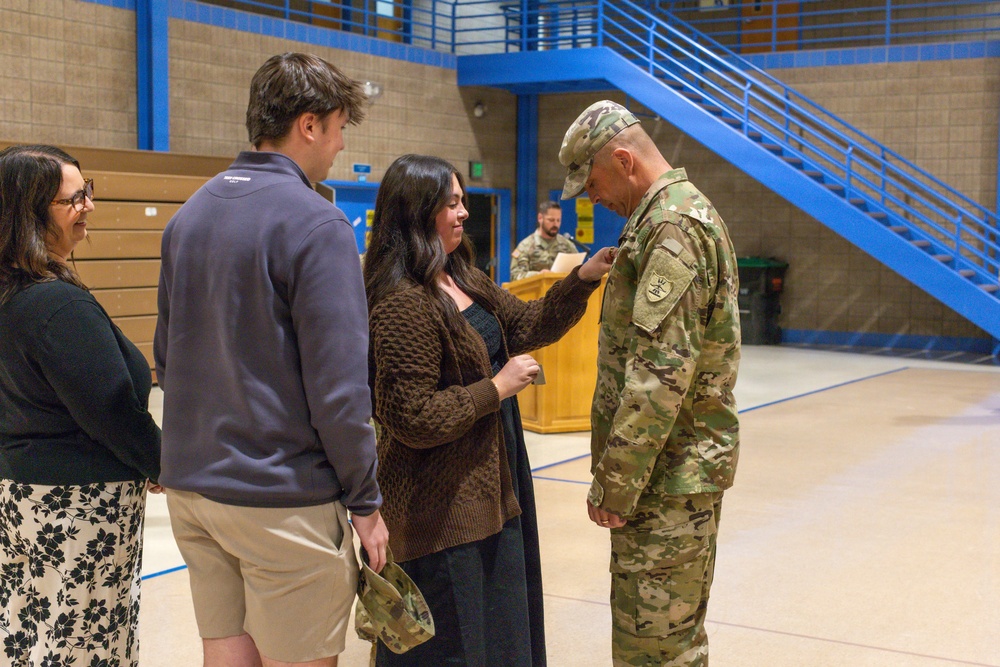 A military promotion or award ceremony taking place inside what appears to be a gymnasium or indoor sports facility. A woman in a brown cardigan is pinning something onto the uniform of a soldier in OCP (Operational Camouflage Pattern) camouflage, while two other civilians look on. This type of ceremony typically involves family members pinning new rank insignia onto a service member.