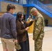 A military promotion or award ceremony taking place inside what appears to be a gymnasium or indoor sports facility. A woman in a brown cardigan is pinning something onto the uniform of a soldier in OCP (Operational Camouflage Pattern) camouflage, while two other civilians look on. This type of ceremony typically involves family members pinning new rank insignia onto a service member.