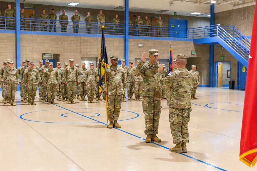 A formal U.S. Army military ceremony is taking place inside a gymnasium or indoor drill hall with blue-accented architecture. A soldier in the center is raising his right hand, suggesting an oath of office or promotion ceremony, while a flag bearer holds a unit guidon nearby. Rows of soldiers in OCP (Operational Camouflage Pattern) uniforms stand in formation, with additional personnel observing from an upper-level mezzanine balcony.
