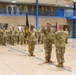 A formal U.S. Army military ceremony is taking place inside a gymnasium or indoor drill hall with blue-accented architecture. A soldier in the center is raising his right hand, suggesting an oath of office or promotion ceremony, while a flag bearer holds a unit guidon nearby. Rows of soldiers in OCP (Operational Camouflage Pattern) uniforms stand in formation, with additional personnel observing from an upper-level mezzanine balcony.