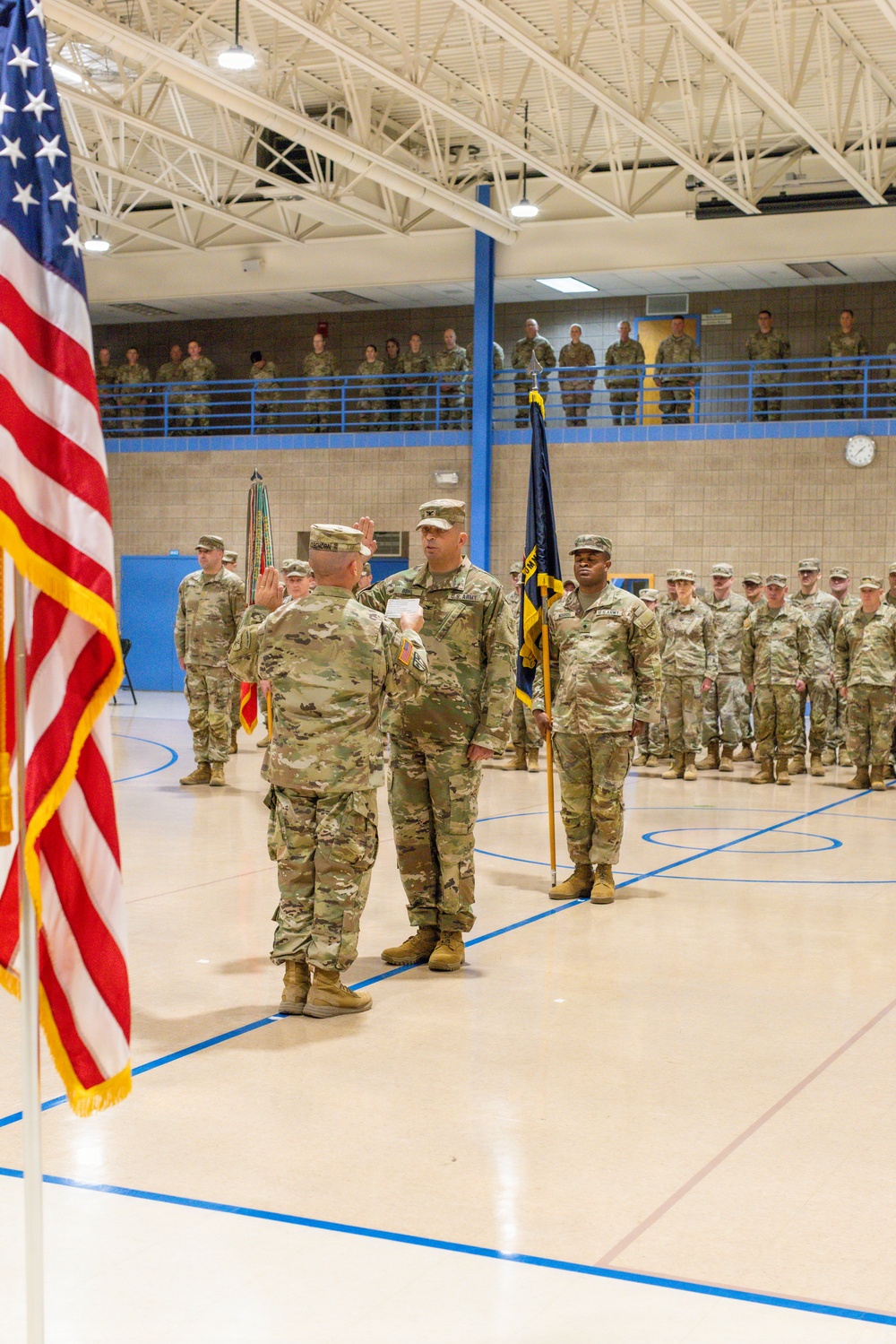 A U.S. Army military ceremony is taking place inside a gymnasium or indoor drill hall. A soldier is administering an oath or conducting a formal ceremony, with one soldier raising their right hand while facing an officiating officer. Troops are formed in ranks on the gymnasium floor and along an upper-level balcony as observers.