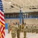 A U.S. Army military ceremony is taking place inside a gymnasium or indoor drill hall. A soldier is administering an oath or conducting a formal ceremony, with one soldier raising their right hand while facing an officiating officer. Troops are formed in ranks on the gymnasium floor and along an upper-level balcony as observers.