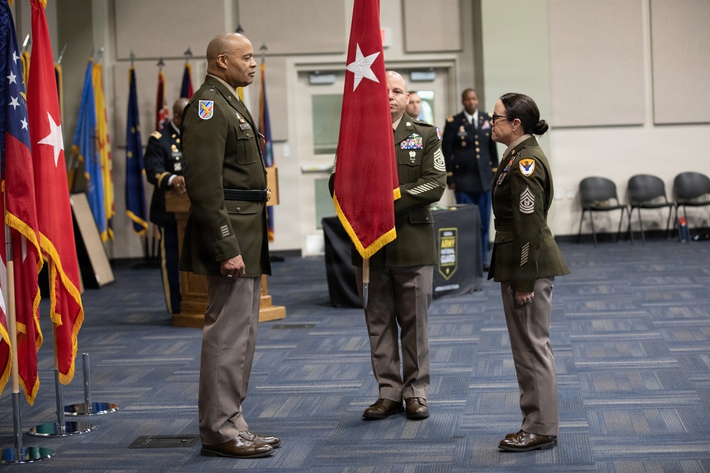 A formal U.S. Army military ceremony is taking place indoors, likely a change of command or promotion ceremony. Three soldiers in Army Green Service Uniforms (Class A) are positioned at the center, with a general officer flag bearer holding a red two-star flag between two senior non-commissioned officers facing each other. Additional military personnel and multiple flags are visible in the background.
