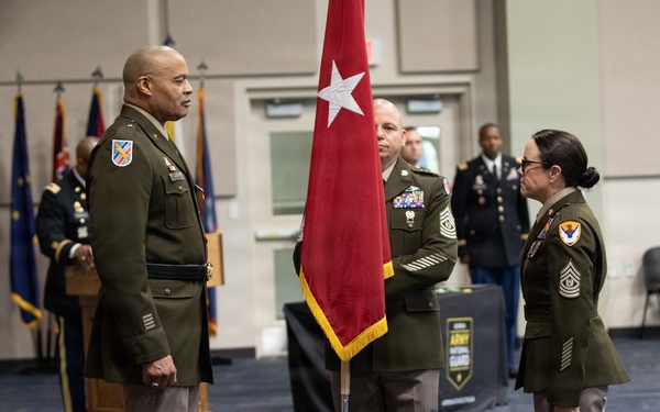 A formal U.S. Army military ceremony is taking place indoors, likely a change of command or promotion ceremony. Three soldiers in Army Green Service Uniforms (Class A) are positioned at the center, with a general officer flag bearer holding a red two-star flag between two senior non-commissioned officers facing each other. Additional military personnel and multiple flags are visible in the background.