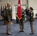 A formal U.S. Army military ceremony is taking place indoors, likely a change of command or promotion ceremony. Three soldiers in Army Green Service Uniforms (Class A) are positioned at the center, with a general officer flag bearer holding a red two-star flag between two senior non-commissioned officers facing each other. Additional military personnel and multiple flags are visible in the background.
