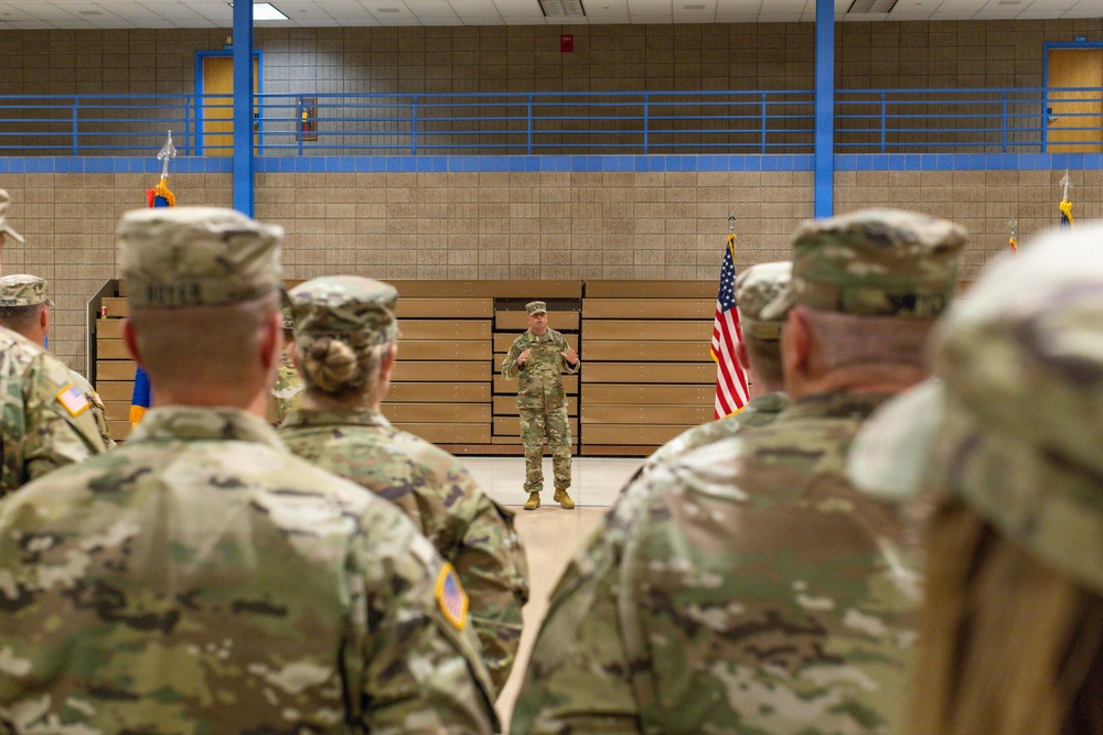 A military briefing or formation is taking place inside what appears to be a gymnasium or indoor drill hall. A single soldier stands at the front addressing a group of uniformed personnel who have their backs to the camera. An American flag and several unit guidons are visible in the background.