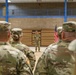 A military briefing or formation is taking place inside what appears to be a gymnasium or indoor drill hall. A single soldier stands at the front addressing a group of uniformed personnel who have their backs to the camera. An American flag and several unit guidons are visible in the background.